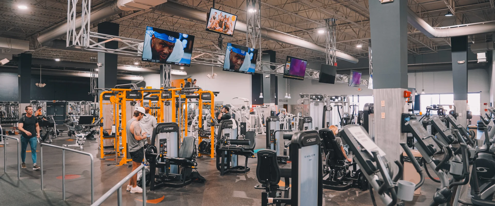 Members working out in the resistance machines area at Gold’s Gym Farragut, Tennessee. The gym features a variety of Cybex strength-training machines and a functional training zone with bright orange Synergy equipment. Overhead, mounted TVs display sports and fitness content, while the industrial-style ceiling and modern lighting enhance the open and dynamic workout environment.