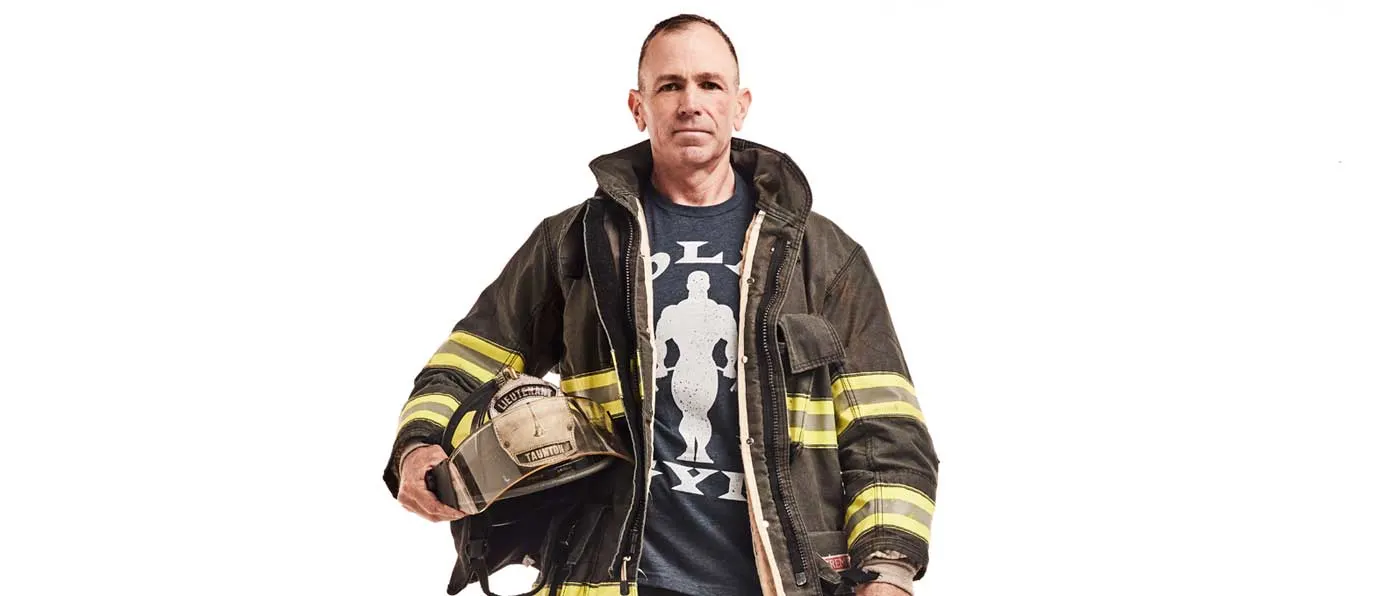 A middle-aged man wearing a firefighter's uniform and holding a helmet stands against a plain white background.