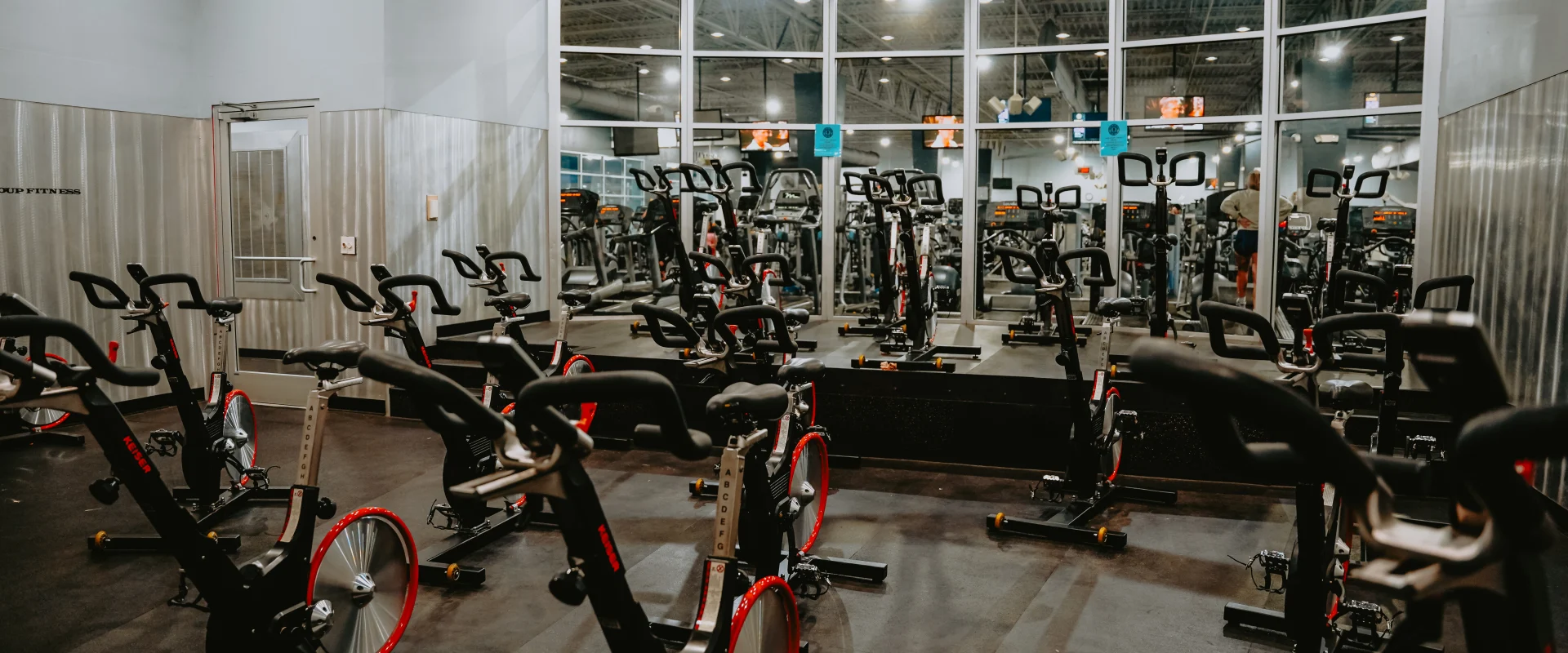 Group cycle studio at Gold’s Gym Tennessee, Lee Highway, featuring rows of stationary bikes arranged on a dark rubber floor. The space includes a raised platform for the instructor and large windows in the background providing a view of the gym floor. The room is illuminated by overhead ceiling lighting, creating a bright environment for cycling classes.