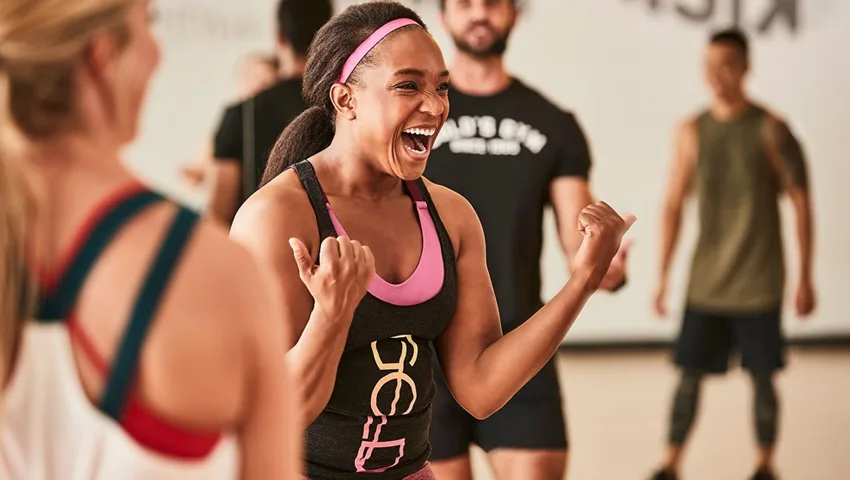 A woman in a pink sports bra and shorts is enthusiastically celebrating with clenched fists, surrounded by other people in workout attire in what appears to be a fitness studio or gym.
