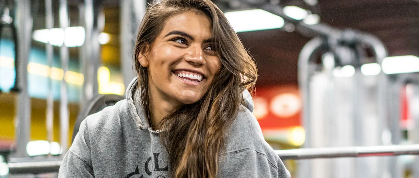 A young woman with long, brown hair is smiling brightly, standing in front of a blurred background with colorful lights.