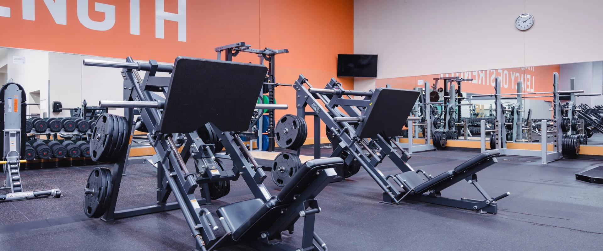 A gym with two leg press machines in the foreground. An orange wall displays the phrase "BUILD YOUR STRENGTH." Mirrors line the back wall, reflecting exercise equipment.