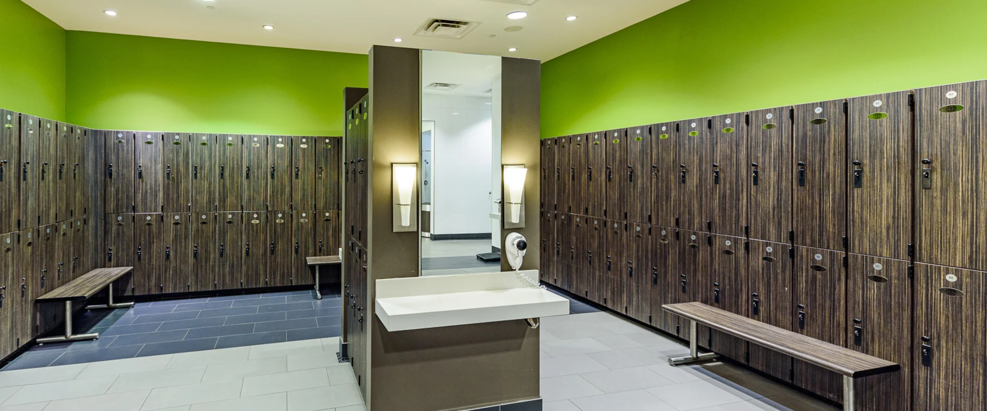 Locker room at Gold’s Gym Hollywood, featuring rows of dark wood-finished lockers with built-in locks lining the walls. Benches with metal legs are placed throughout the space. A central vanity area includes a white sink, a mounted mirror, and a wall-mounted hairdryer. Two modern wall sconces provide additional lighting. The flooring consists of large light and dark tiles, contrasting with the bright green accent walls. Recessed ceiling lights illuminate the area.
