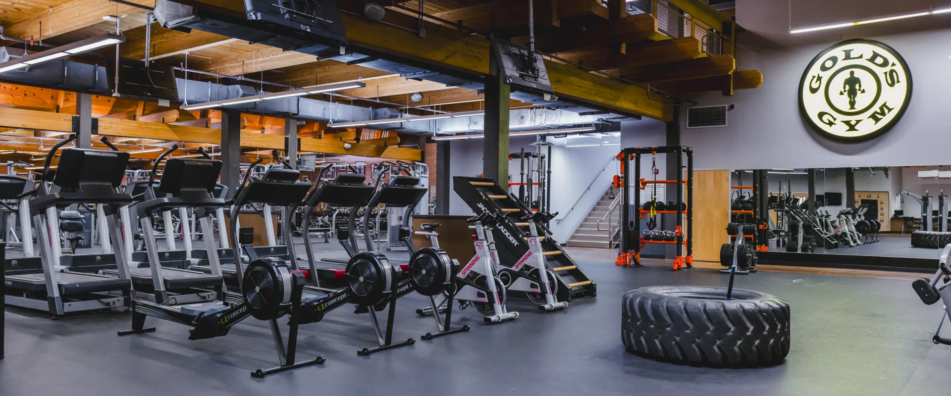 A functional training area at Gold's Gym featuring treadmills, rowers, bikes, a sled, a large tire, and vibrant orange workout racks under a wooden ceiling.
