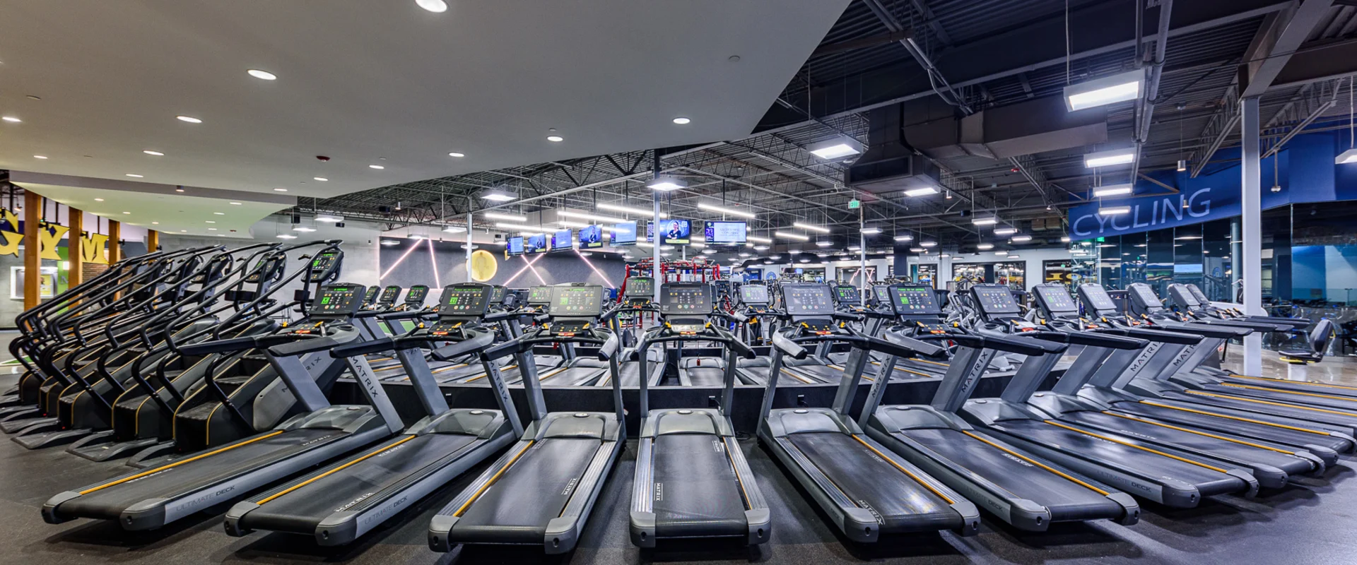 A vast lineup of treadmills and elliptical machines stretches across the floor, each equipped with a digital screen. Bright overhead lighting reflects off polished black flooring, with the "Cycling" room visible in the background.