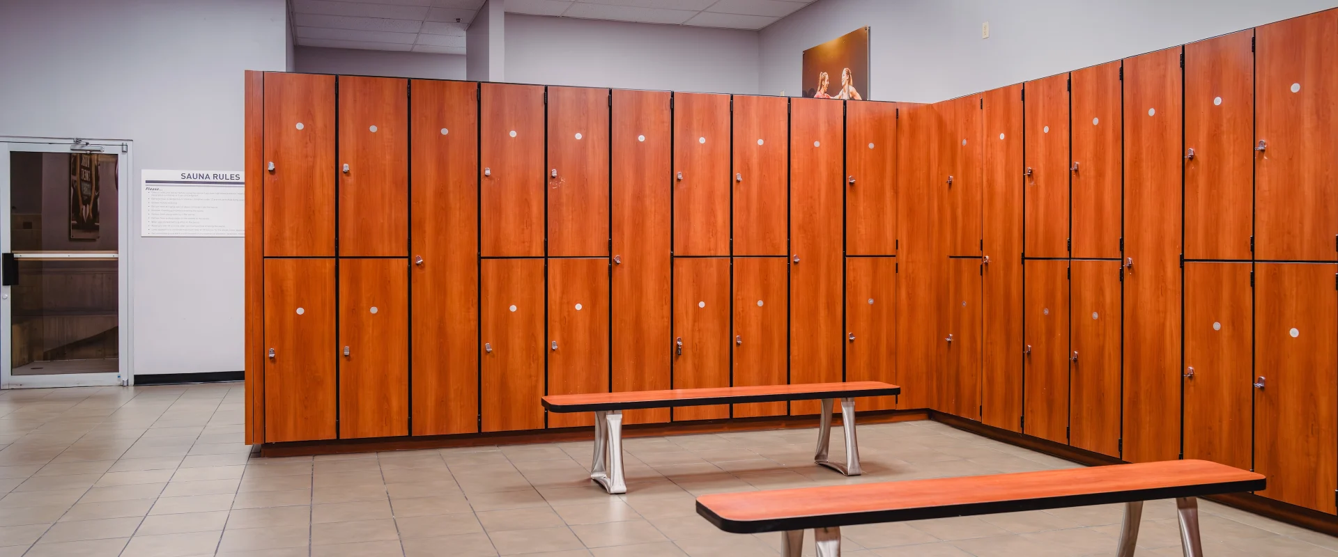 Locker room at Gold’s Gym in Austin (South Central), featuring rows of wooden lockers with metal handles and secure locks. The space includes tiled flooring, sturdy wooden benches with metal legs, and bright overhead lighting. A sign displaying sauna rules is visible near the entrance, providing easy access to additional amenities.