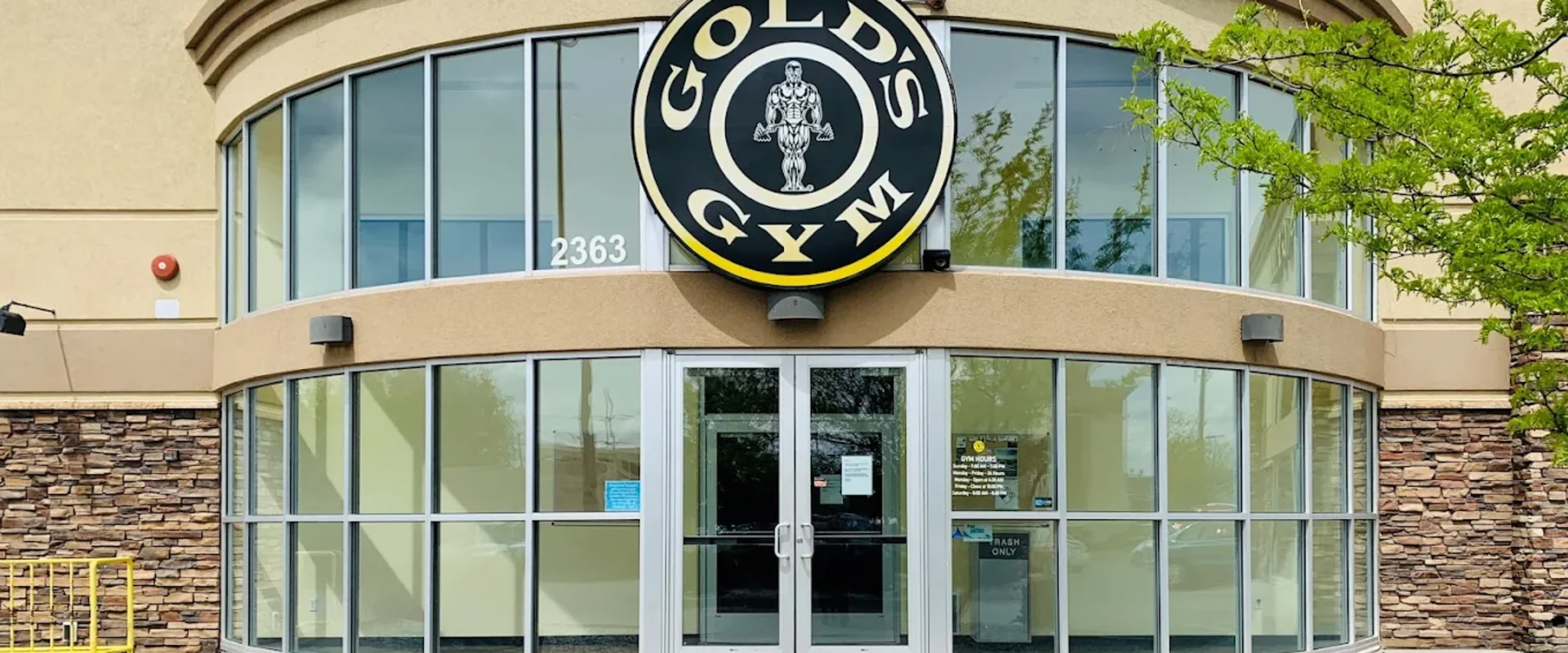 Front entrance of Gold’s Gym with curved glass windows, tan walls, stone accents, and the Gold’s Gym logo above the doors.