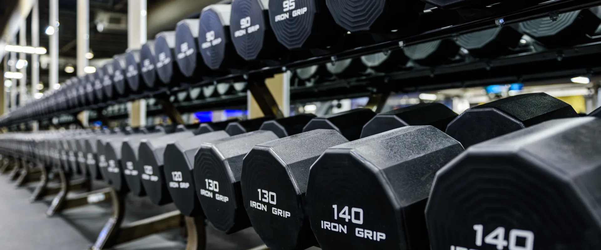 Close-up of the dumbbell rack at Gold’s Gym Thousand Oaks, showing a row of heavy dumbbells neatly arranged on a black metal rack. The dumbbells have a hexagonal shape with weight markings in white text. The reflection in the mirror reveals more gym equipment in the background. Overhead LED lighting casts a bright glow on the space.