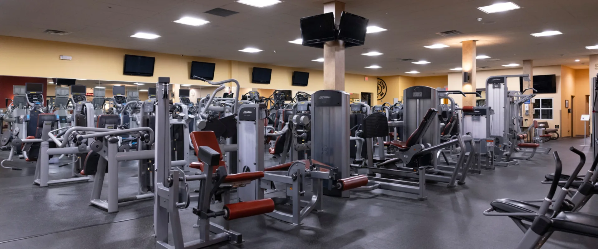 Strength equipment area at Gold’s Gym in East Northport, New York, featuring Life Fitness selectorized machines for targeted muscle training, including leg curl, chest press, and shoulder press machines. The space is organized with rubber flooring, mirrored walls, and multiple overhead TVs. Bright ceiling lights enhance visibility, with additional cardio equipment visible in the background.