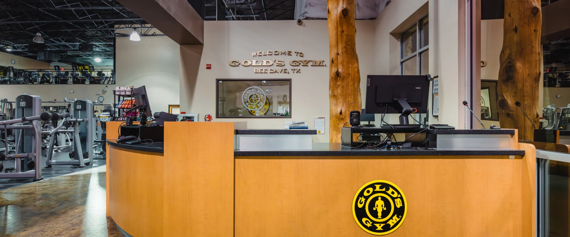Front desk area at Gold's Gym Austin (Bee Caves) featuring a curved wooden reception counter with the Gold's Gym logo. The desk is equipped with a computer. Behind the desk, a window to an office displays the gym’s logo. The space includes natural wood accents and overlooks the main workout floor.