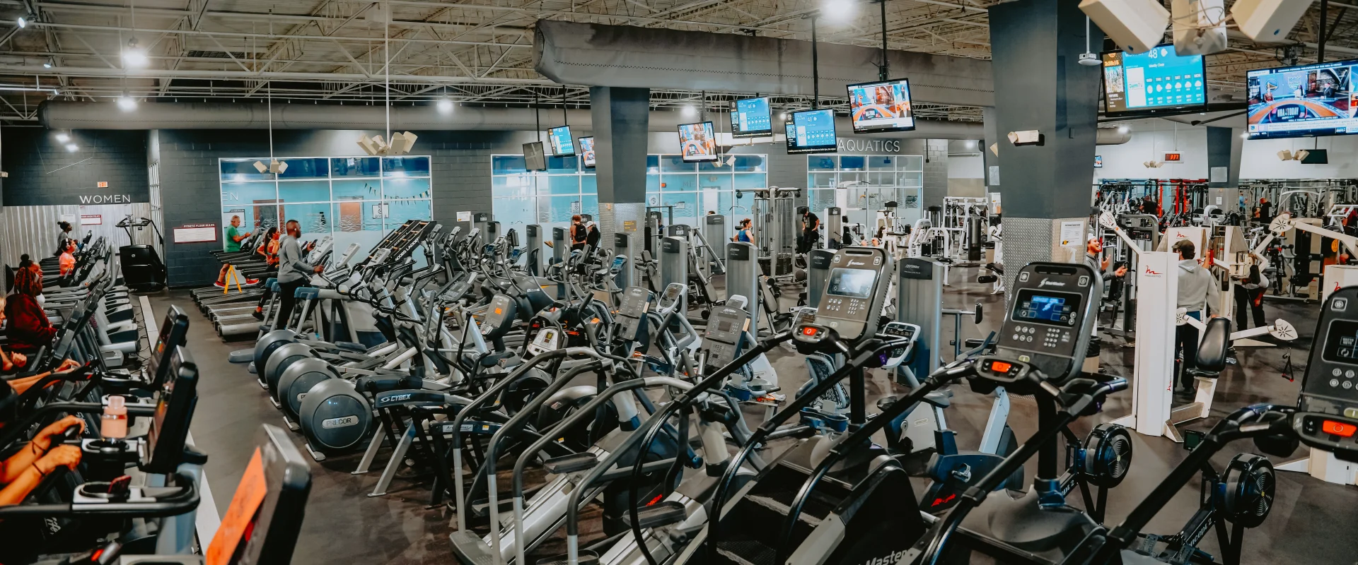 Cardio equipment area at Gold’s Gym Tennessee, Lee Highway, featuring rows of treadmills, elliptical machines, and stair-climbing machines on dark rubber flooring. The space includes a high ceiling with exposed beams, hanging light fixtures, and multiple mounted screens displaying workout stats and entertainment. A view of the aquatics center and machine weight area is visible in the background.