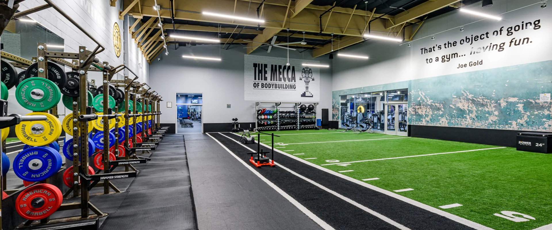 The turf training area at Gold’s Gym Cerritos features a large stretch of artificial turf marked with running lanes, positioned next to a row of squat racks with color-coded bumper plates. A wall mural reads ‘The Mecca of Bodybuilding,’ and a motivational quote by Joe Gold is displayed on an adjacent wall. The area includes sleds, plyometric boxes, and other functional training equipment.