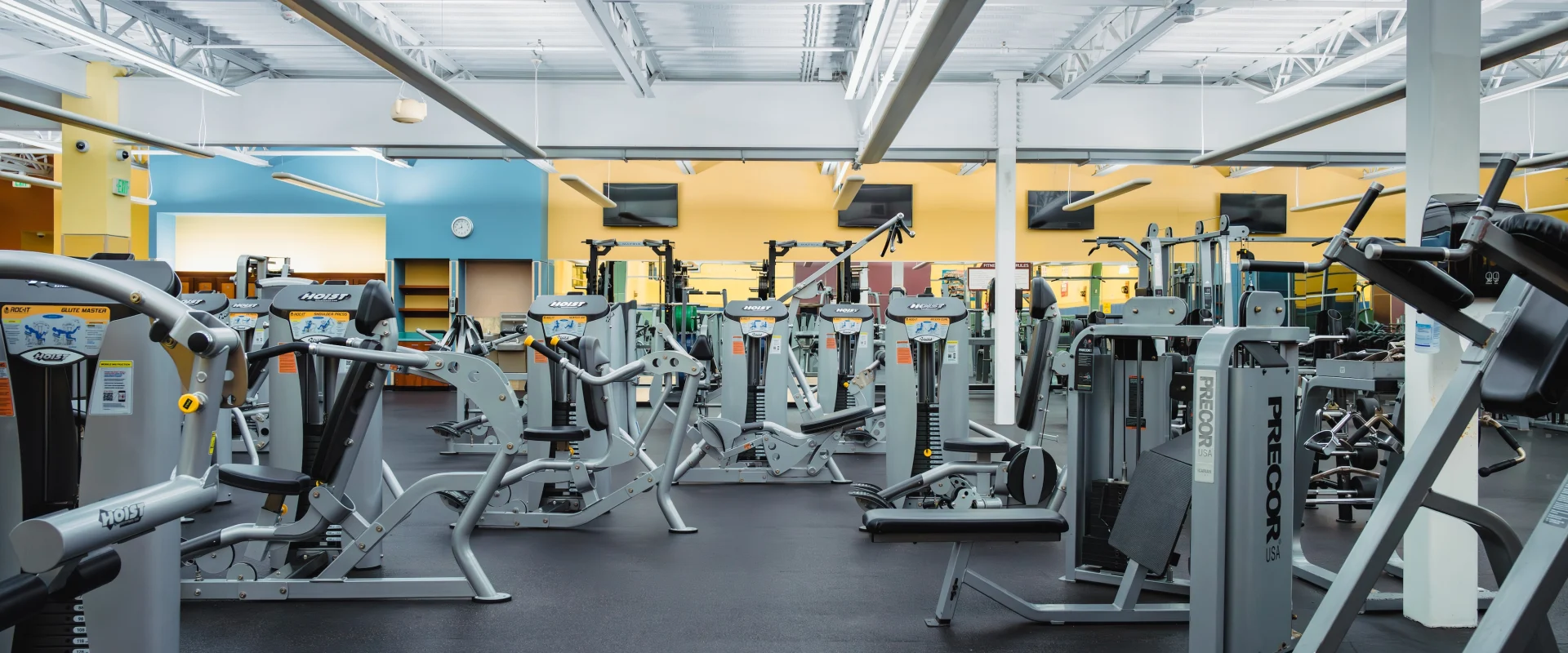 Spacious gym with various exercise machines, including treadmills and weight machines, under bright overhead lighting and a clean, organized layout.