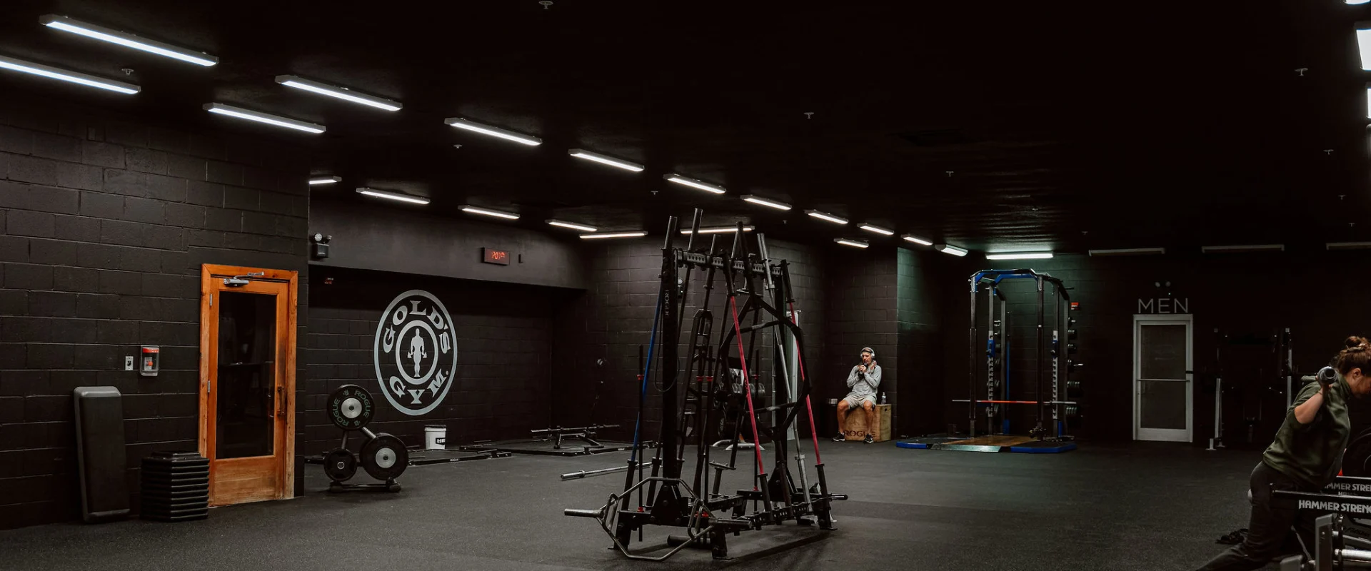 Functional training area at Gold’s Gym, Chapman, Tennessee, featuring resistance bands, free weights, and functional training racks positioned in the center of the room. The black walls and ceiling, paired with linear overhead lighting, create a focused and modern training environment. A Gold’s Gym logo is displayed prominently on one wall, while a small group of members engages in activities around the space. Adjacent equipment like squat racks and benches are visible, enhancing the versatility of the functional training zone.