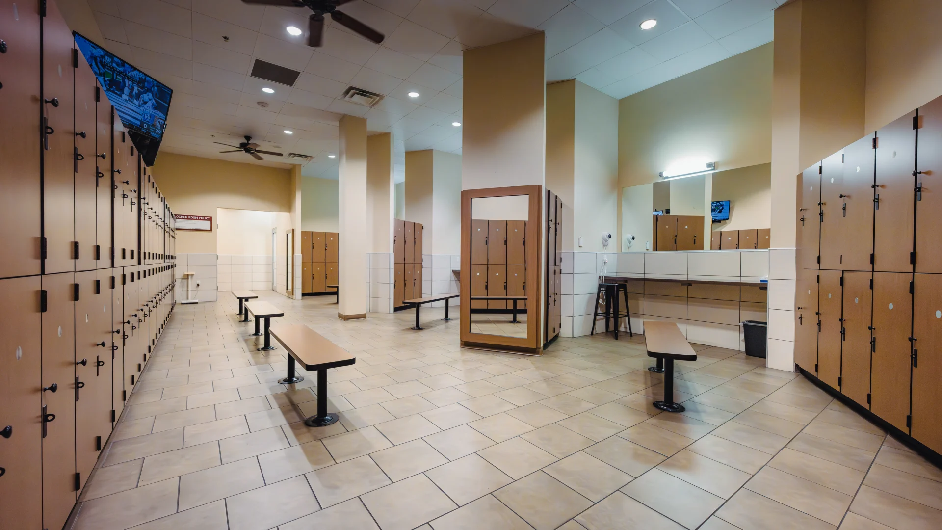 Locker room at Gold’s Gym Austin (Anderson Arbor) with beige tiled flooring, wooden lockers lining the walls, and benches in the center. A full-length mirror, a grooming area with sinks, mirrors, and hair dryers, and a wall-mounted TV are visible. Ceiling fans and recessed lighting illuminate the space.