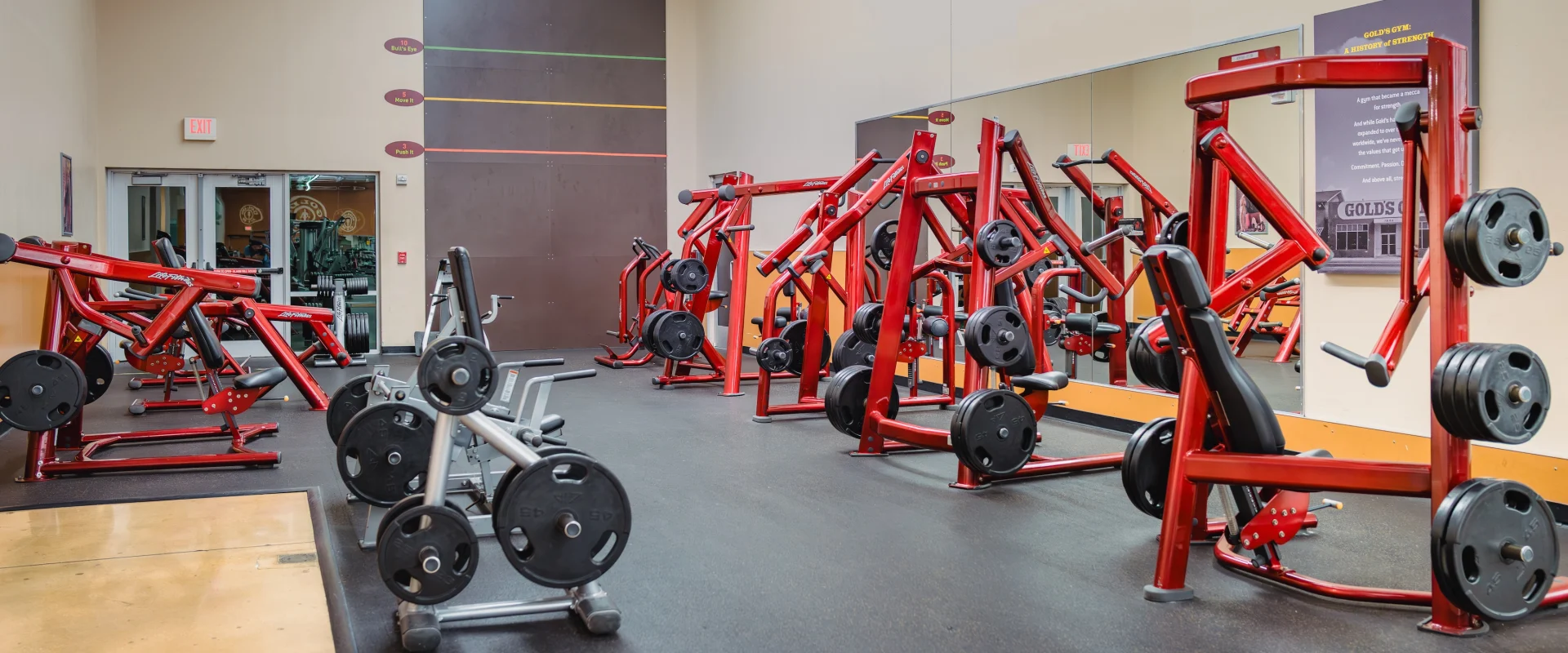Spacious gym interior with red weight machines, black rubber flooring, and mirrors reflecting equipment. Bright lighting creates an energetic atmosphere.