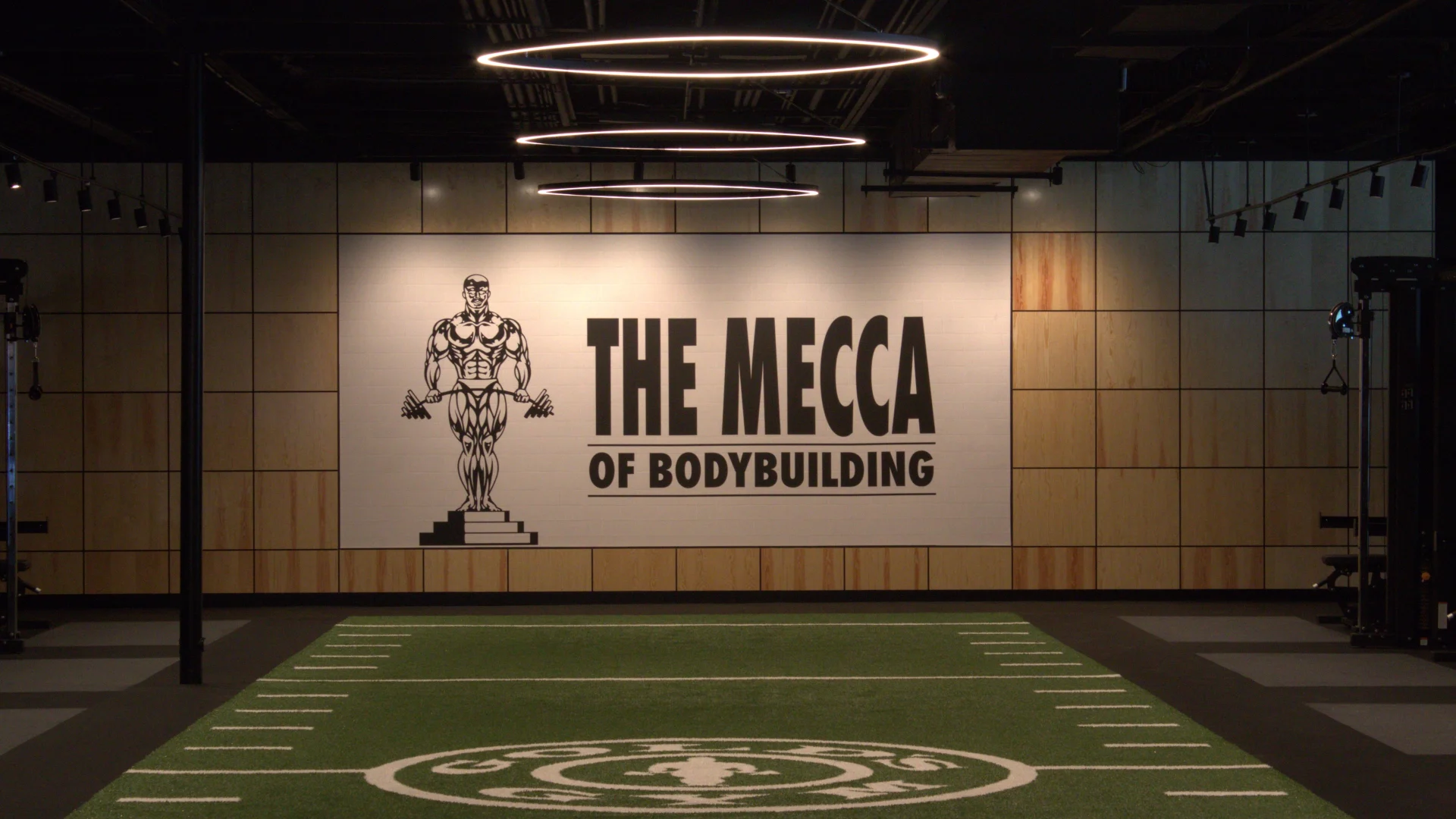 Indoor turf field at Gold's Gym Austin, Burnet Rd with Gold's Gym logo printed on the turf and “The Mecca of Bodybuilding” mural prominently displayed on the far wall.