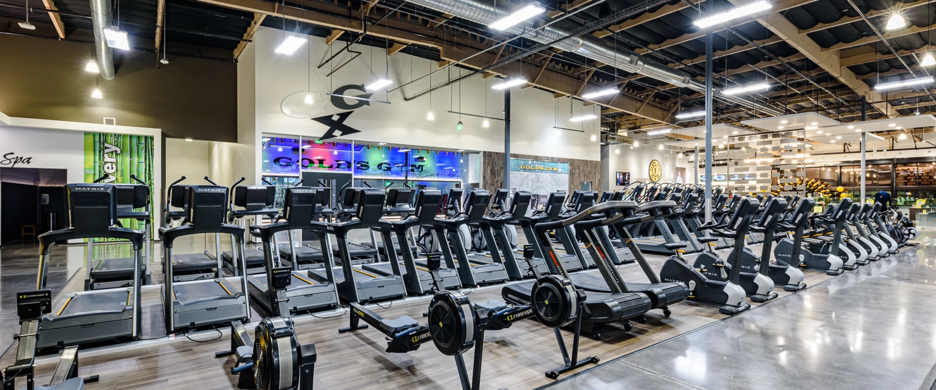 The cardio area at Gold’s Gym Cerritos features rows of treadmills, rowing machines, and stationary bikes lined up on wood-style flooring. Overhead TVs are mounted on metal poles, displaying various channels.