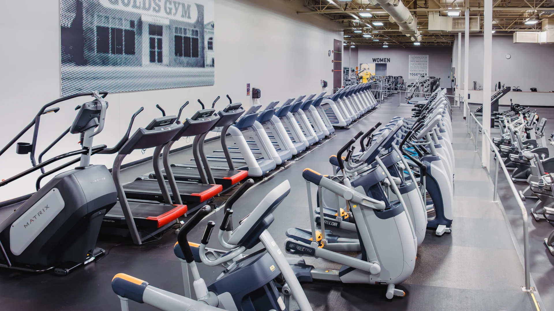 Cardio area at Gold's Gym Waco featuring rows of treadmills, ellipticals, and stationary bikes on a black rubber floor, with a vintage Gold's Gym Venice photo mural on the wall and high ceilings with industrial-style lighting.