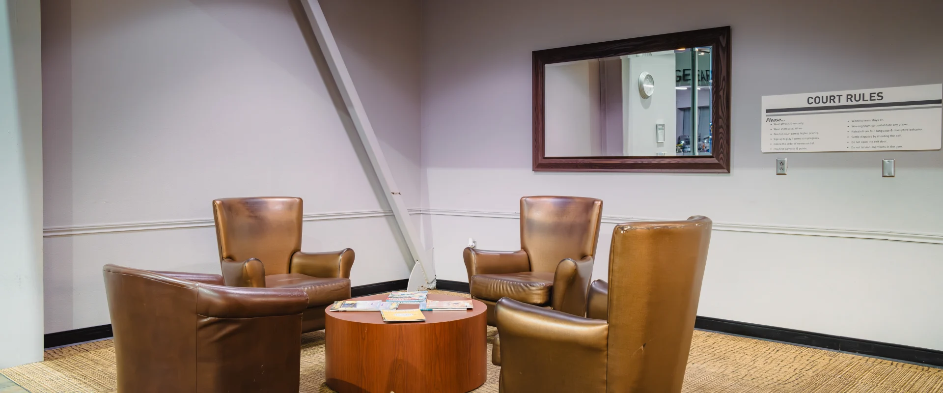 Lounge area in Gold's Gym Waco featuring three brown leather chairs around a round wooden table, set on a tan carpet, with a large wall mirror and court rules signage in the background.