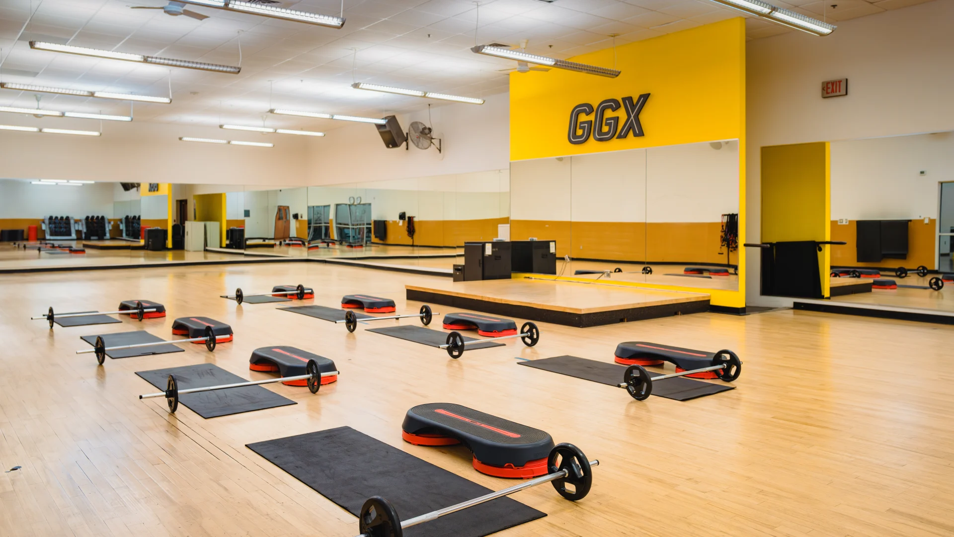 Group exercise room at Gold's Gym in Austin (Hesters Crossing) featuring wood flooring, mirrored walls, and yellow accents. Equipment, including barbells, mats, and steps, is evenly arranged for a class setup. A raised platform at the front serves as an instructor stage. The room is well-lit with overhead lights and ceiling fans.