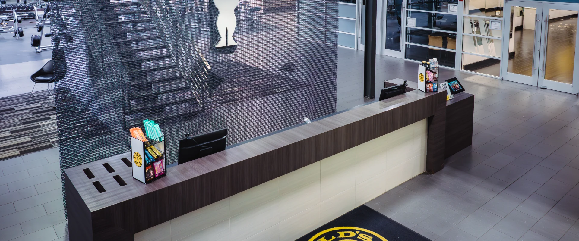 The front desk area at Gold's Gym Oklahoma City (Moore), featuring a modern design with a silhouette of the Gold's Gym weightlifter logo on a textured chain-link wall, a prominent staircase in the background, a kiosk for check-ins, a display of snacks on the counter, and a Gold's Gym-branded floor mat.