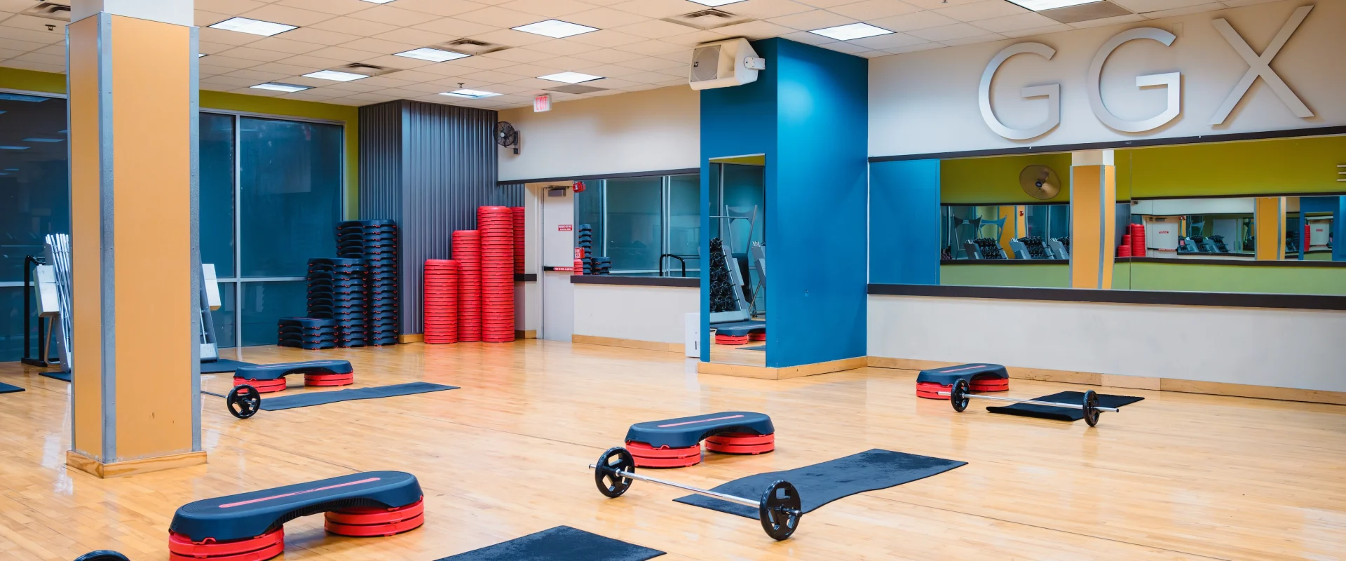 Spacious gym studio with wood flooring, exercise mats, weights, and steps neatly arranged. Reflective mirrors and "GGX" on the wall create a clean, energetic atmosphere.