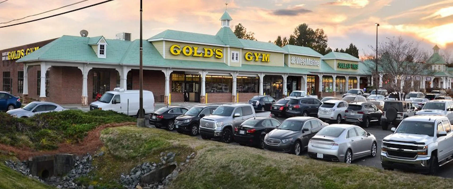 Exterior of Gold’s Gym Lake Norman with yellow signage and green roof in shopping plaza at sunset.