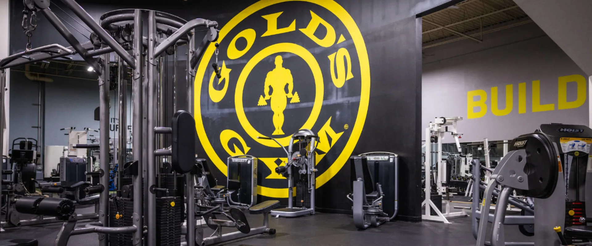A multi-station cable machine sits in the foreground, surrounded by various selectorized strength machines. The Gold’s Gym logo dominates the black wall in the background, with large yellow lettering. The flooring is rubber, and mirrors reflect additional equipment. A plate-loaded machine is partially visible on the right.