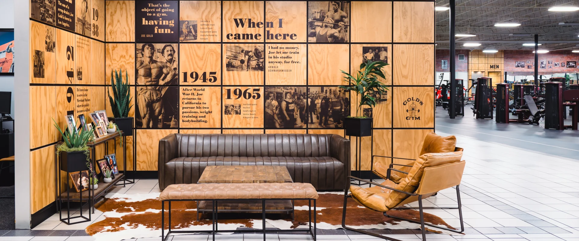Stylish gym lounge with a leather sofa, tan chair, wood panel wall with historical photos and text, potted plants, and a tiled floor. Cozy ambiance.