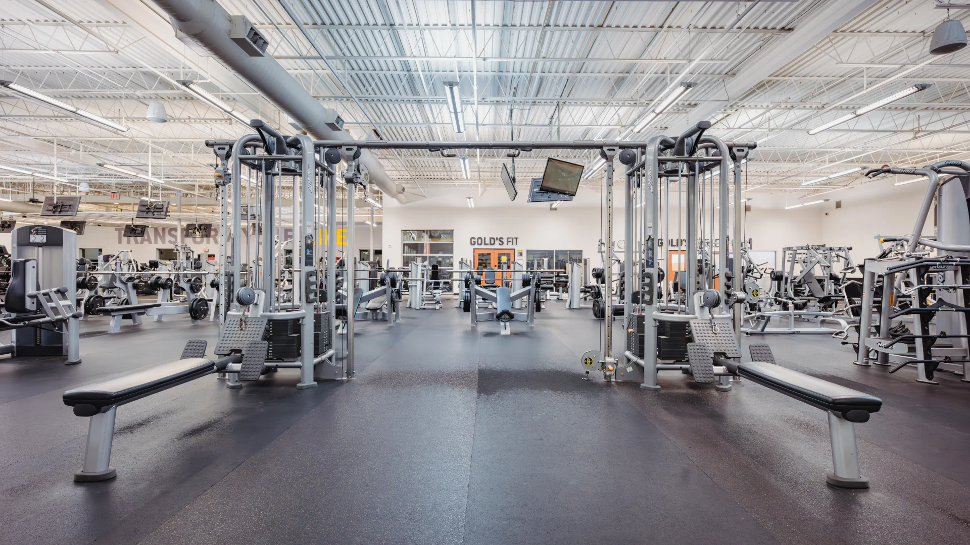 Spacious gym area featuring machine weight equipment arranged in rows on black rubber flooring. Centralized cable machines are flanked by benches and adjustable seats, while additional strength-training machines are visible in the background. The high ceiling with exposed ductwork enhances the open atmosphere, with overhead fluorescent lighting providing even illumination. A "Gold's Fit" sign and windows are visible in the rear wall, adding branding and separation to the space. Monitors hang from the ceiling for entertainment.