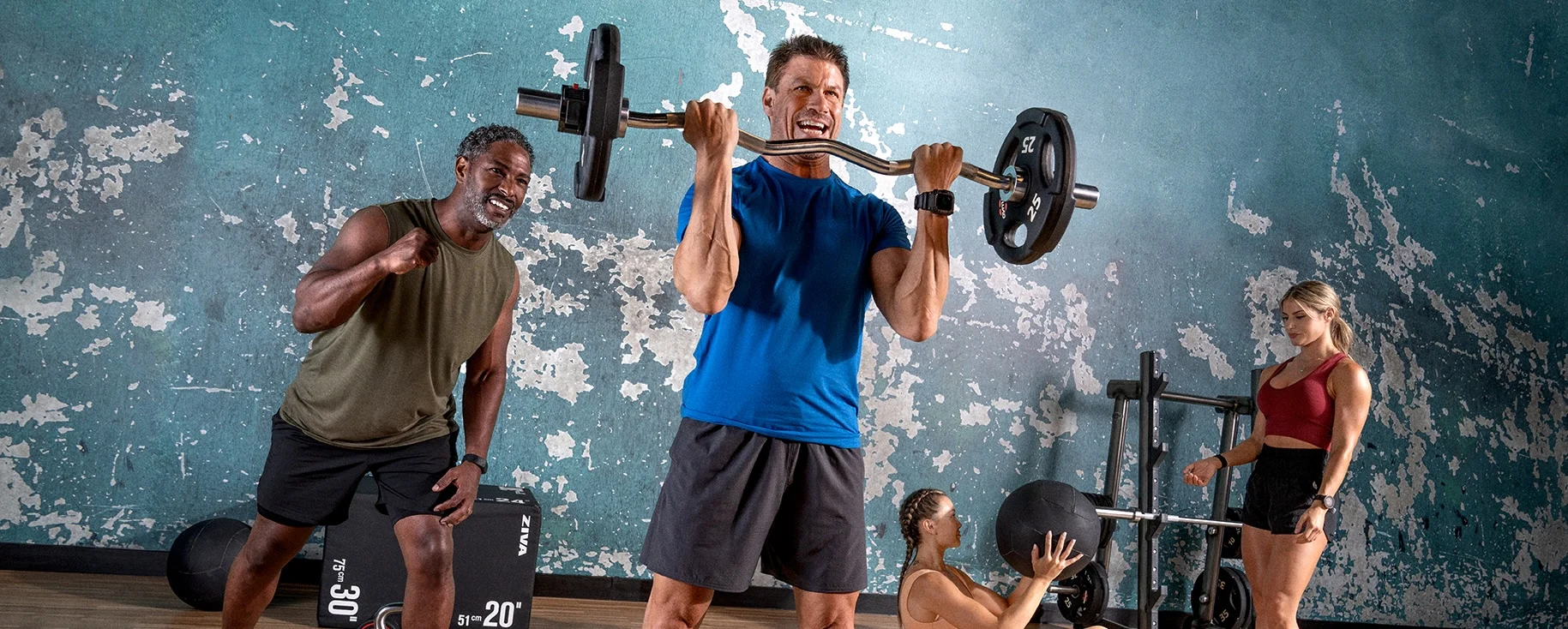 Four people perform beginner strength exercises in a gym; one lifts a barbell, another cheers, while two women train with a medicine ball.