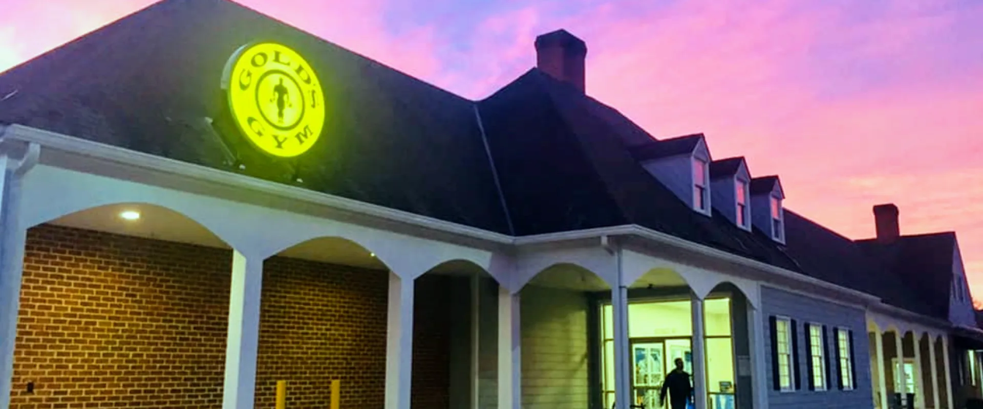 Exterior of Gold’s Gym Gayton Crossing with lit yellow logo on brick building under pink and purple sunset sky.
