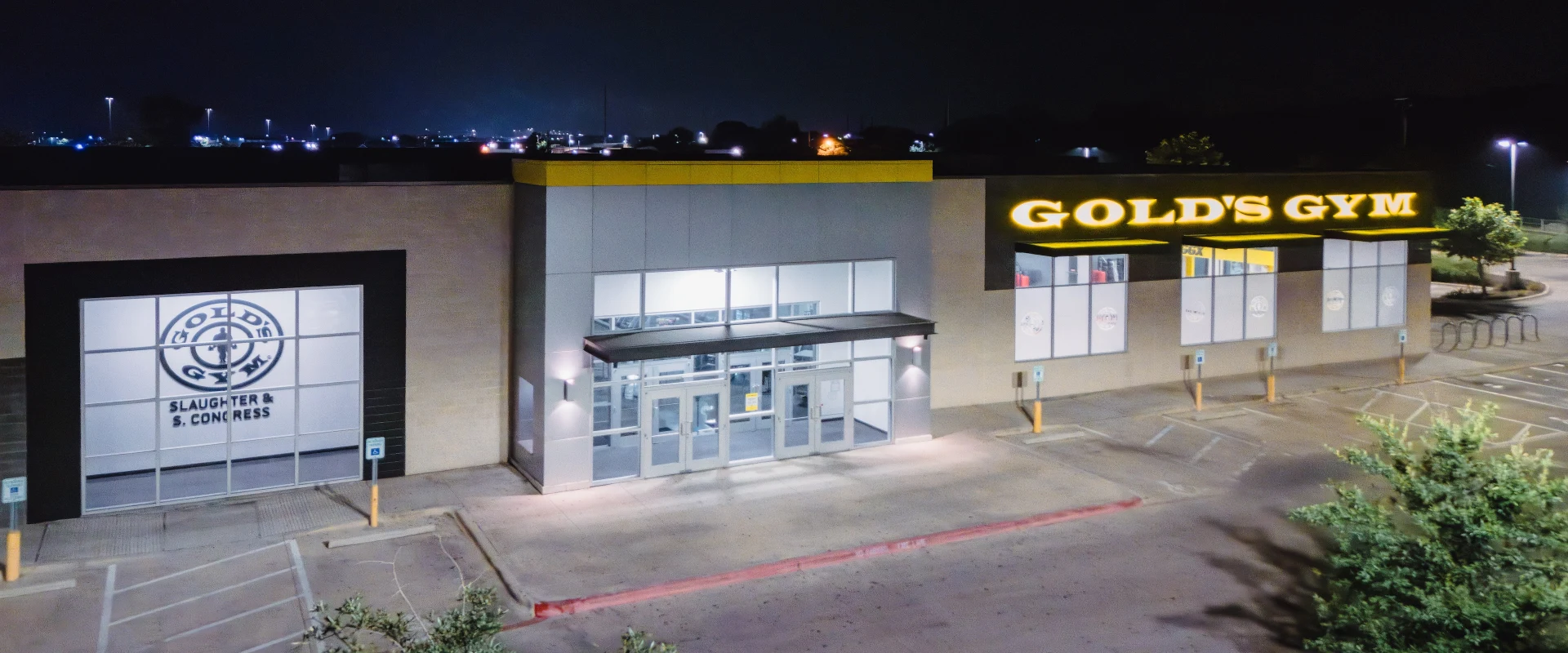 The exterior of Gold's Gym in Texas (Slaughter & South Congress) showcases a modern facade with large glass windows and the iconic Gold's Gym logo illuminated at night. The entrance features double glass doors, while the surrounding parking lot is well-lit for safety.