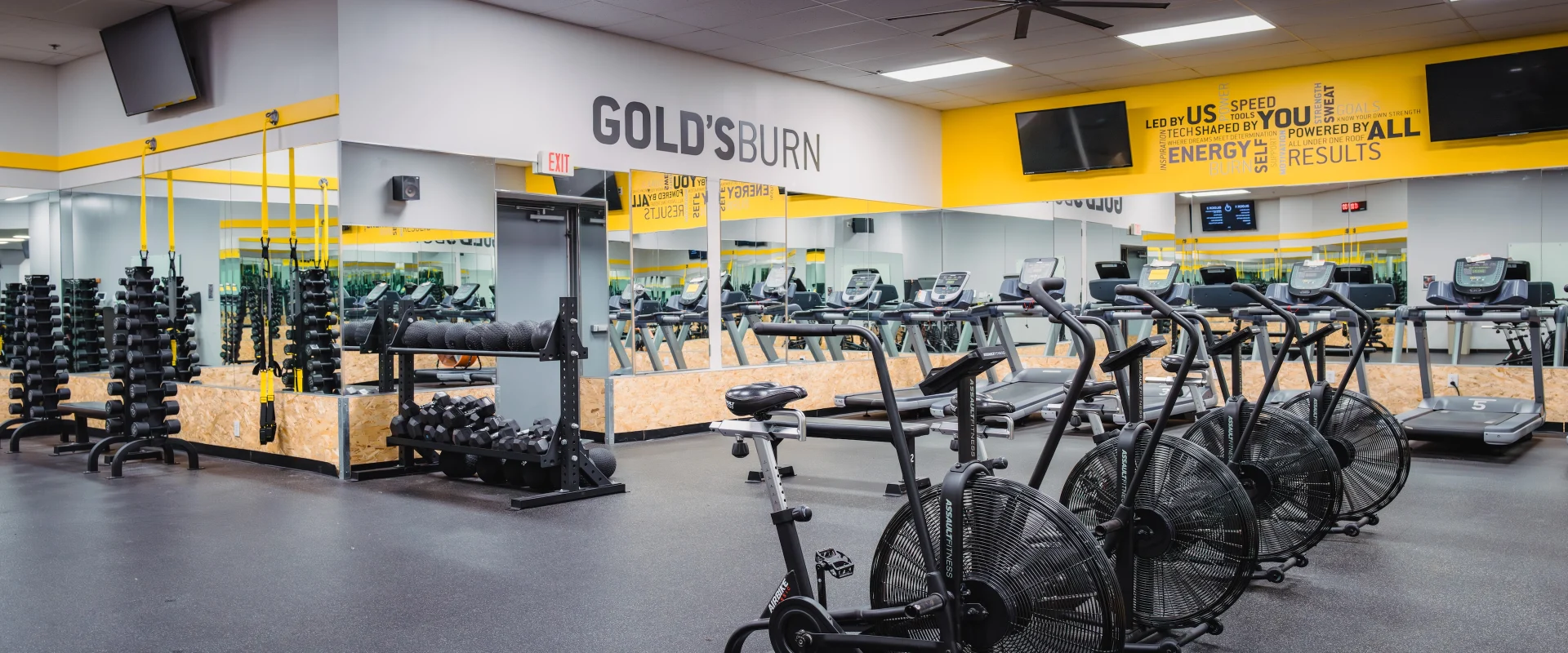 Gold’s Burn studio at Gold’s Gym in Austin (South Central), featuring rows of air bikes, treadmills, and free weights arranged on rubber flooring. The space includes TRX suspension trainers, dumbbell racks, and foam rollers, with mirrored walls enhancing visibility. Bright yellow accent walls with motivational text and mounted TVs create an energetic environment for high-intensity interval training.