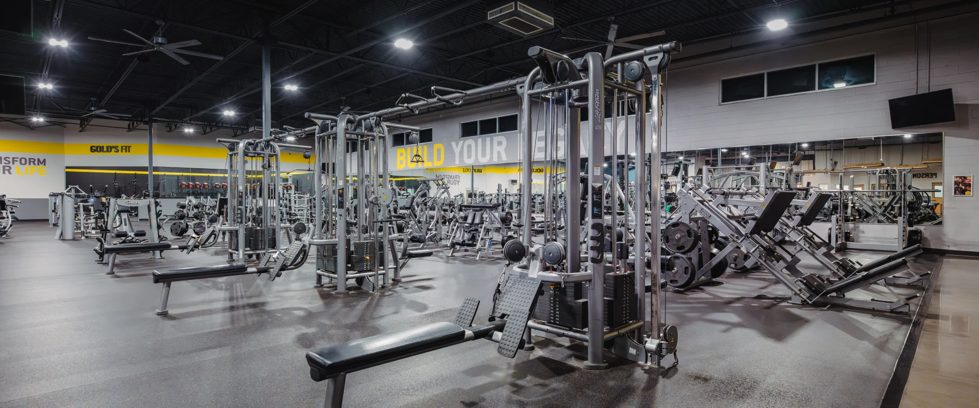 Weight machine area at Gold’s Gym Belterra, featuring various strength-training equipment, adjustable benches, and mirrored walls under high ceilings with bright industrial lighting. Yellow accent walls and a display of motivational phrases like "Build Your Legacy."