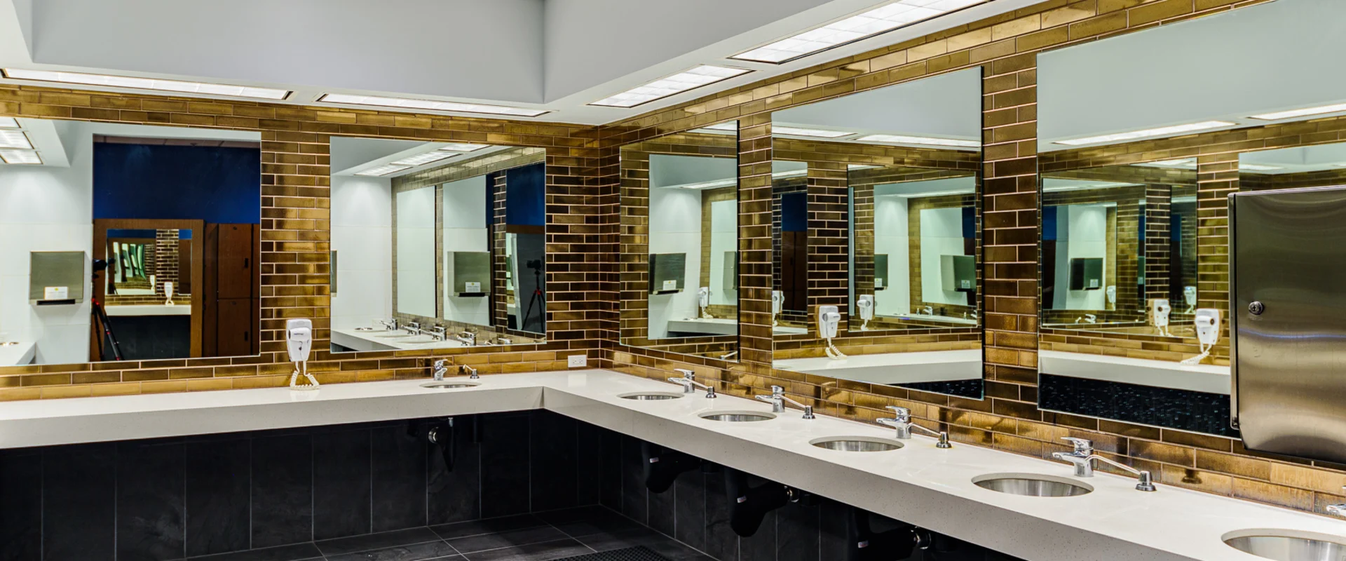 A sleek and modern gym vanity area featuring a long white quartz countertop with multiple stainless steel sinks and chrome faucets. Large mirrors framed by glossy gold subway tiles line the walls, reflecting the bright overhead lighting. Wall-mounted hair dryers and paper towel dispensers provide convenience, while dark tiled flooring and a textured rubber mat enhance the space’s durability and safety.