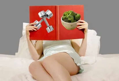 A person sitting on a bed, holding a red book in front of their face and a bowl of broccoli in their hands.