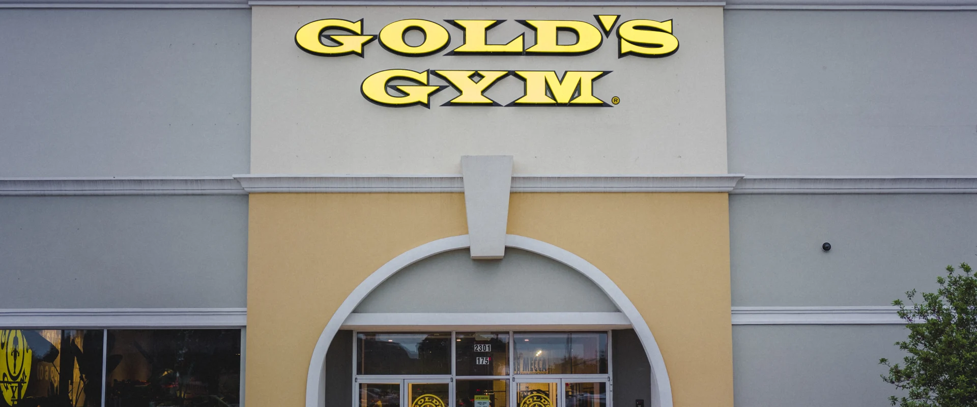 Exterior of Gold’s Gym Flower Mound, showcasing the iconic yellow logo above a glass-front entrance with arched detailing and stone accent building, set against a cloudy sky.