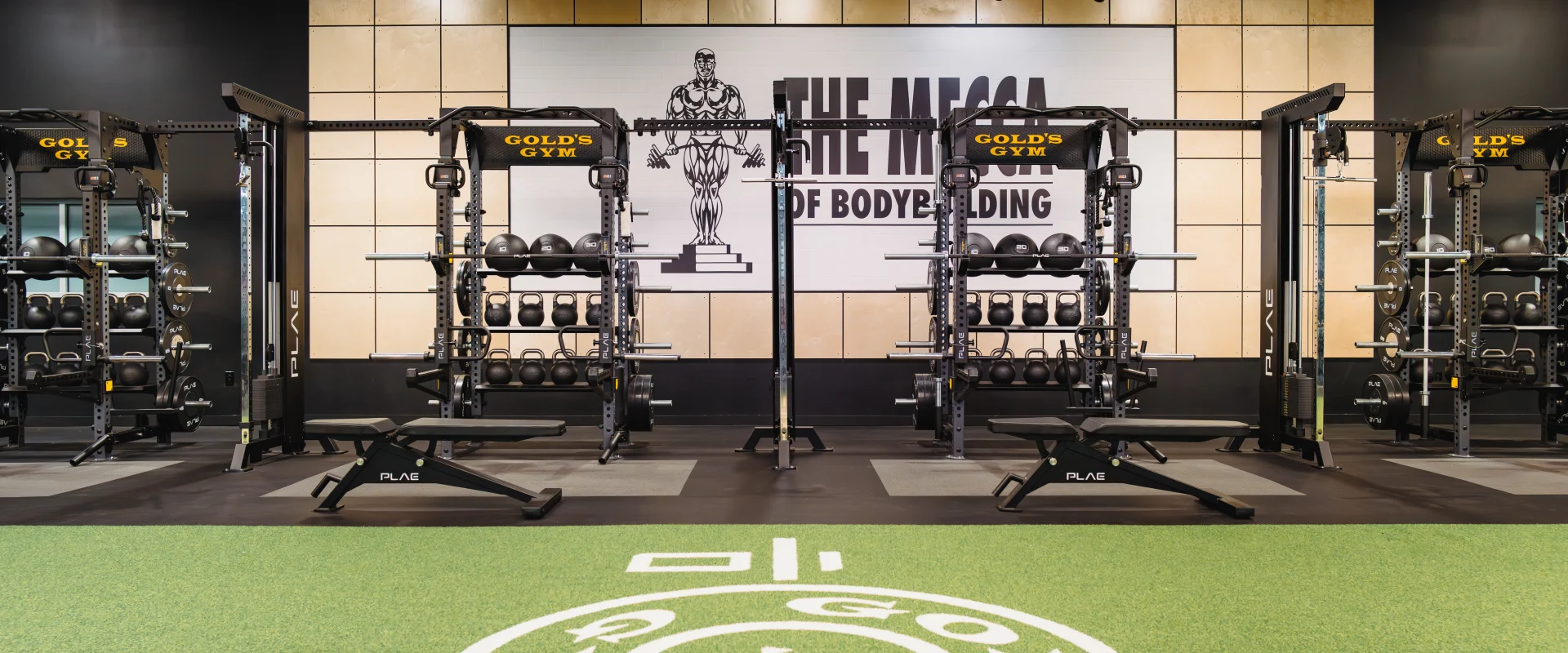 A functional training area at Gold's Gym featuring racks of kettlebells, medicine balls, and weight plates, with 'The Mecca of Bodybuilding' signage on the wall.