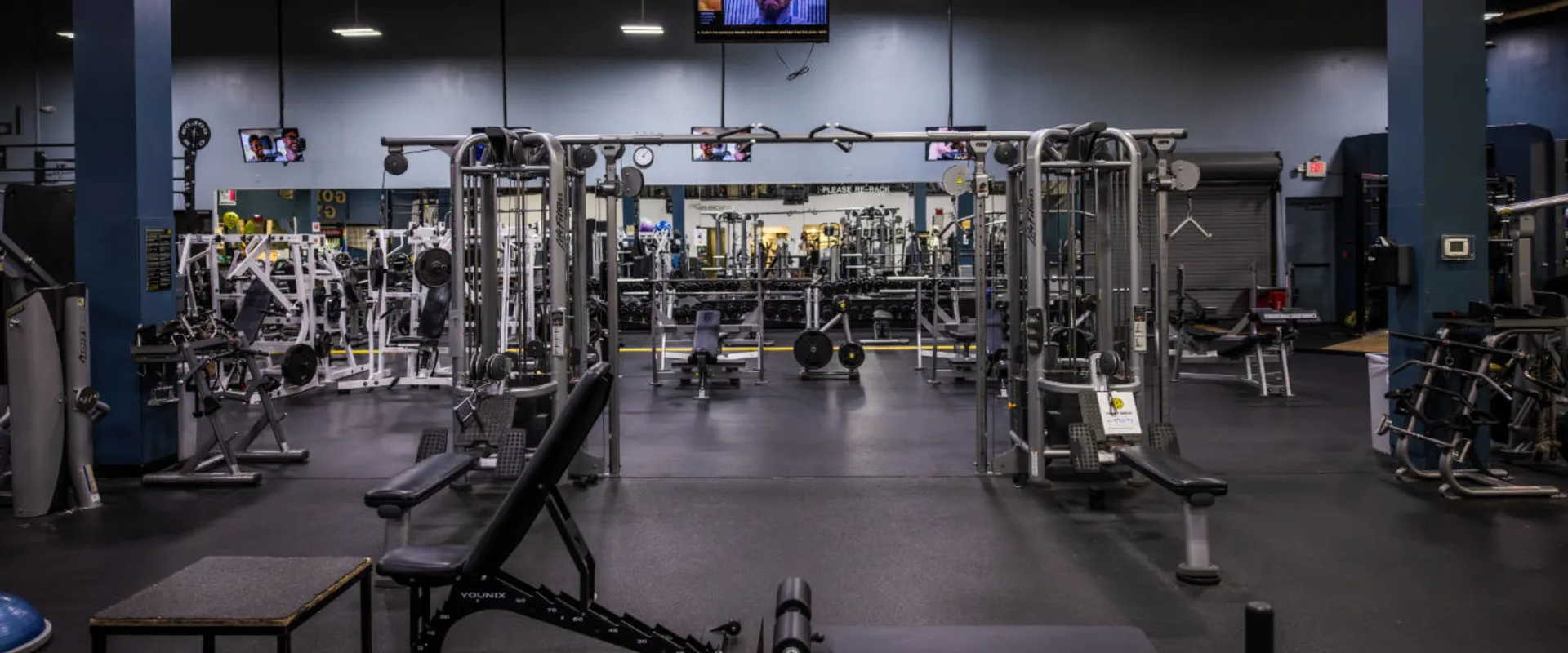 A functional training area at Gold’s Gym in Woodbridge (Lake Ridge) features a large cable crossover machine in the center, surrounded by various strength training equipment. Adjustable benches are positioned nearby, with a squat rack and barbells visible in the background. A mirrored wall reflects the gym space, lined with additional free weights and machines. Several mounted televisions hang from the ceiling, displaying various programs. The dark rubber flooring contrasts with the lighter walls, and overhead lighting illuminates the workout space.