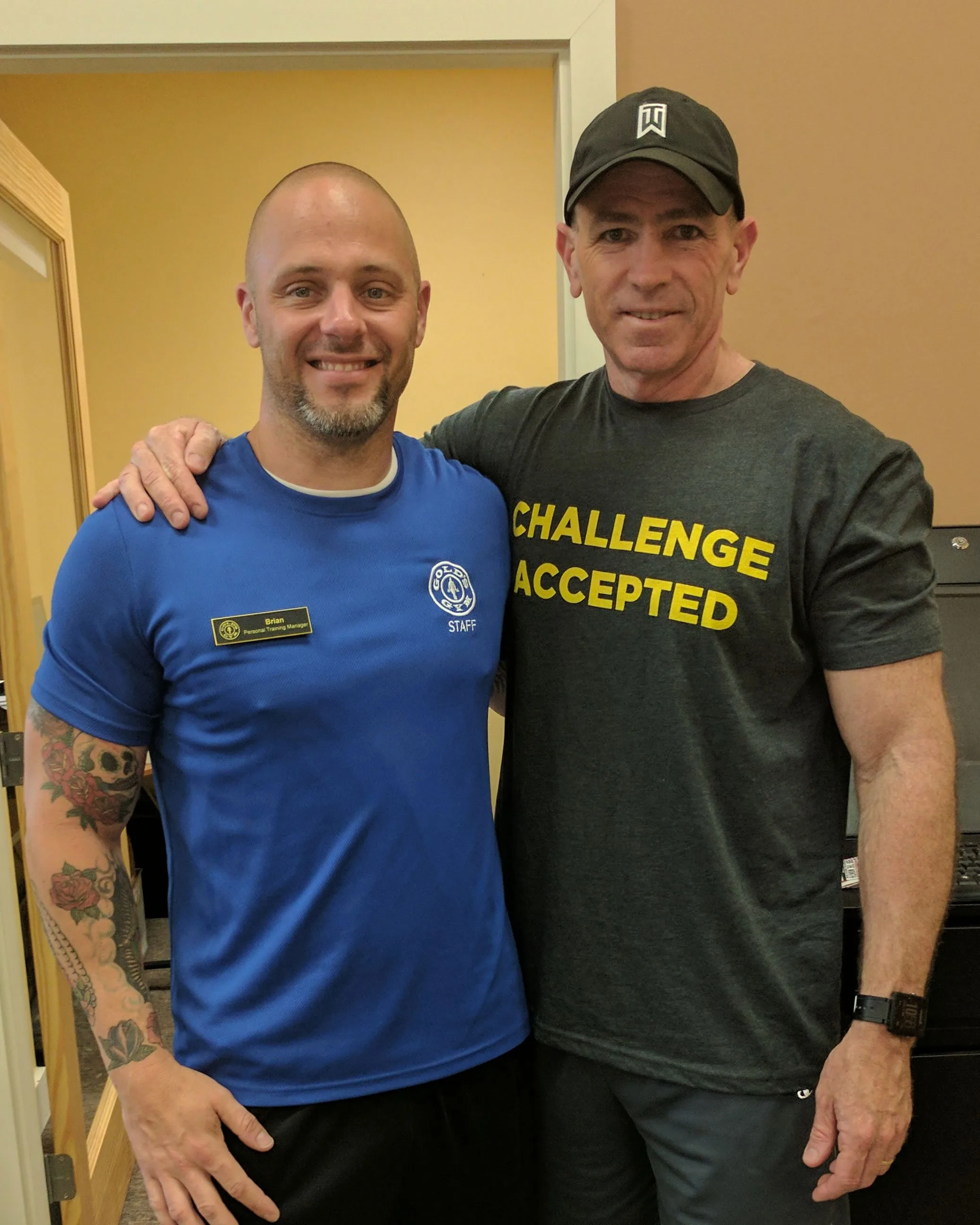 Two men, one wearing a blue shirt and the other a black shirt with the text "Challenge Accepted", are standing together and smiling in what appears to be a room or hallway.