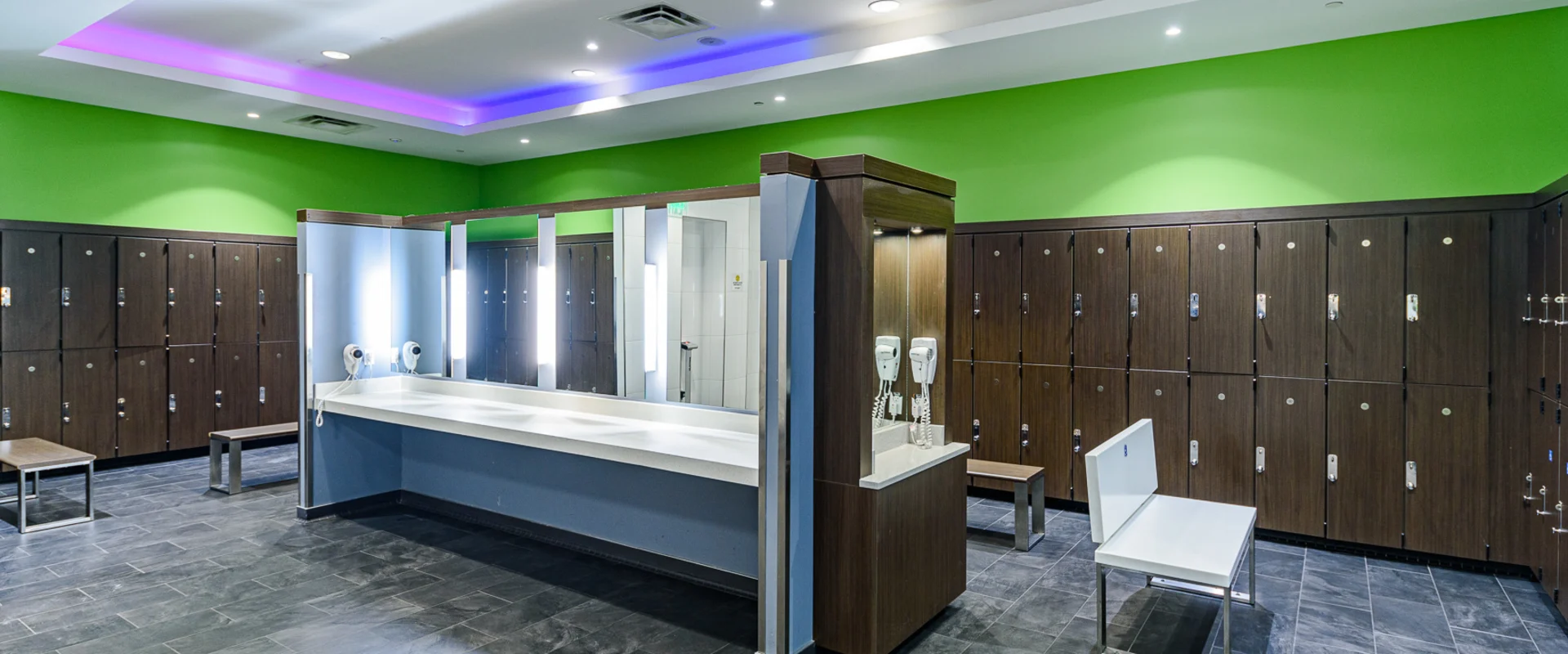 Spacious locker room at Gold's Gym Garden Grove with dark wood lockers, cushioned benches, and a central vanity station. The brightly lit area includes large mirrors, wall-mounted hairdryers, and a modern ceiling with recessed lighting. The floor is covered in dark tiles, contrasting with the vibrant green and blue accent walls.