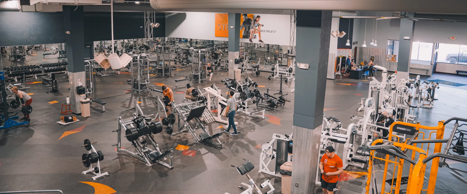 Members exercising in the free weights and resistance training area at a gym in Farragut, Tennessee. The spacious gym floor includes various strength training machines, weight benches, and racks of barbells and dumbbells. A large mirrored wall reflects the equipment and activity, while overhead industrial-style lighting and exposed ducts provide a modern aesthetic. The vibrant orange Synergy360 station adds a pop of color to the room’s neutral tones, emphasizing the gym's functional layout and dynamic atmosphere.