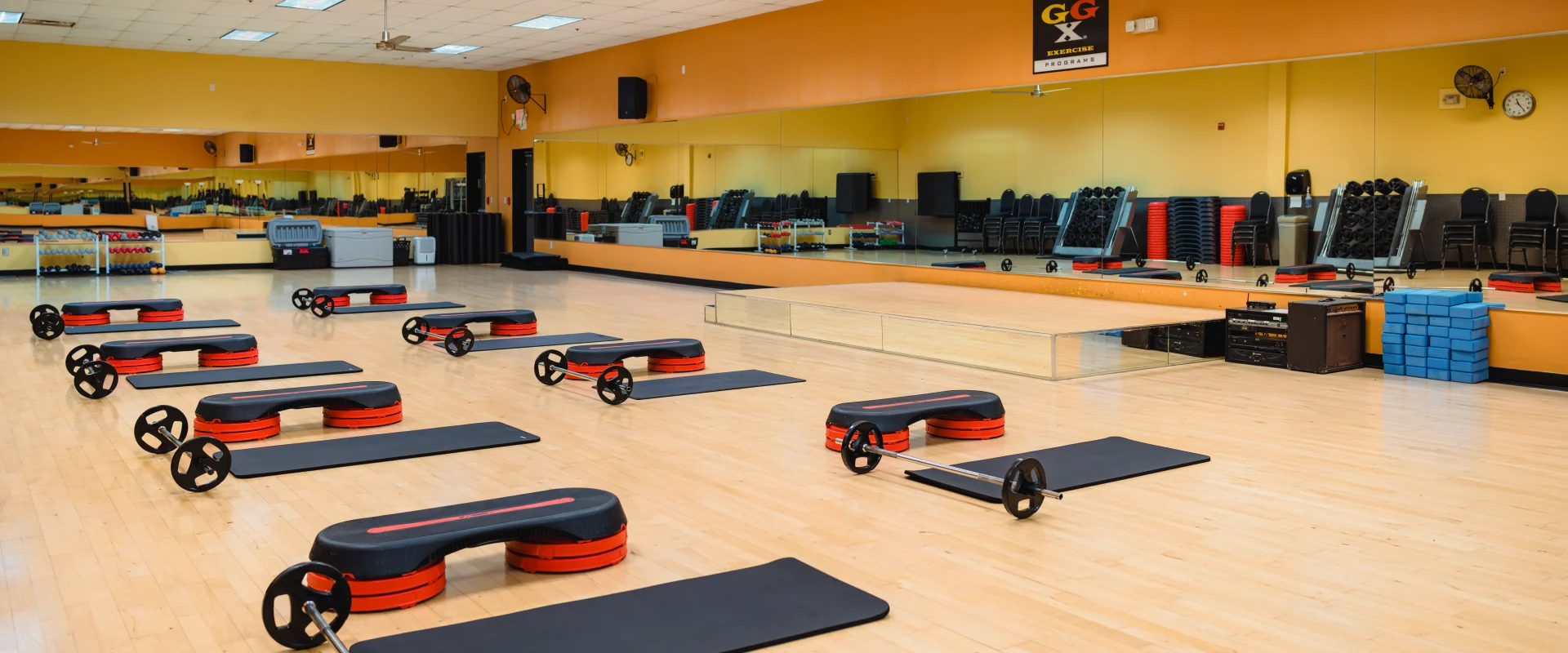 Gold's Gym group exercise studio in Austin (Bee Caves) with step platforms, mats, and barbell weights evenly arranged on a polished wood floor. The room features mirrored walls, yellow and orange accents, and organized equipment racks in the background.