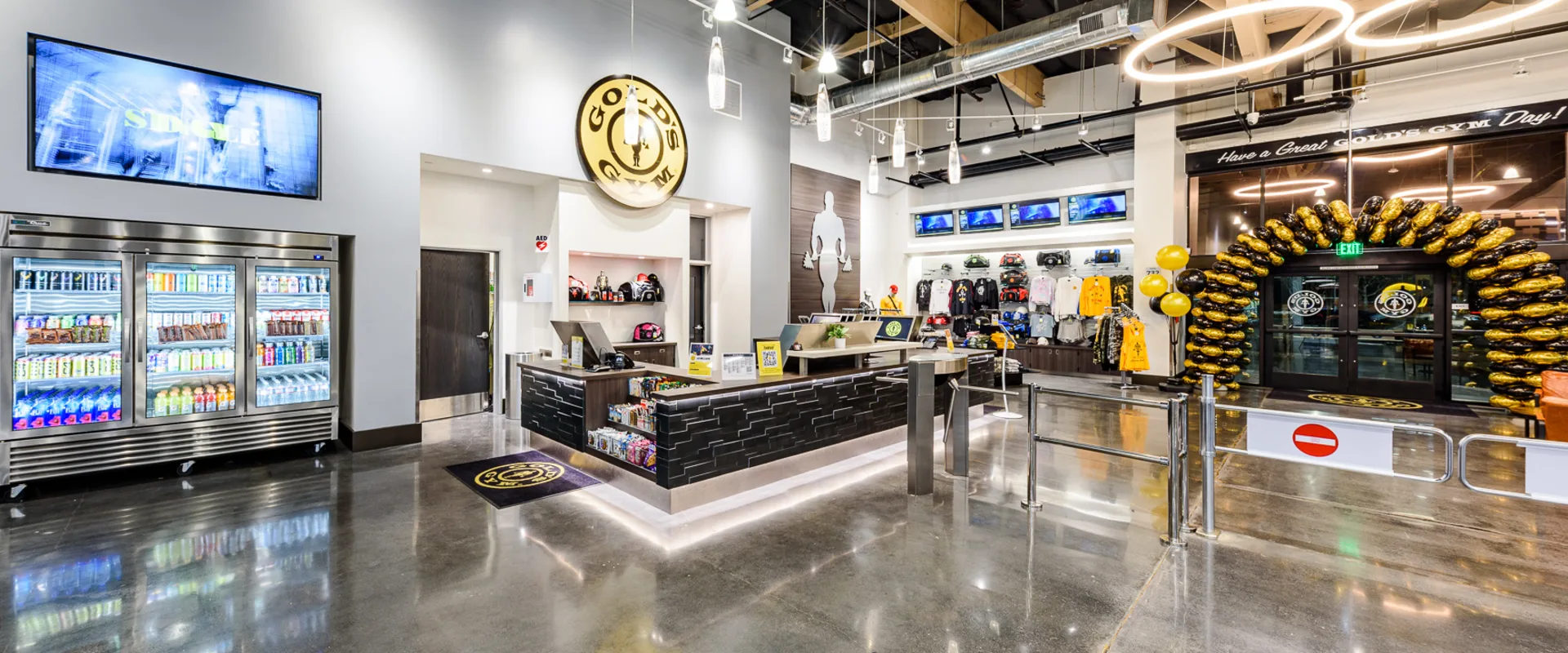 The front desk at Gold’s Gym Cerritos features a sleek black and wood-accented counter with illuminated trim, creating a modern and inviting entrance. Behind the desk, gym essentials such as apparel, bags, and accessories are displayed on white shelving. A Gold’s Gym logo sign is mounted above the reception area, adding a bold brand presence. To the left, a refrigerated display stocked with energy drinks and protein beverages offers convenient refreshments. The entrance is marked by a black-and-gold balloon arch, while polished concrete floors and circular LED light fixtures contribute to the gym’s upscale and contemporary aesthetic.
