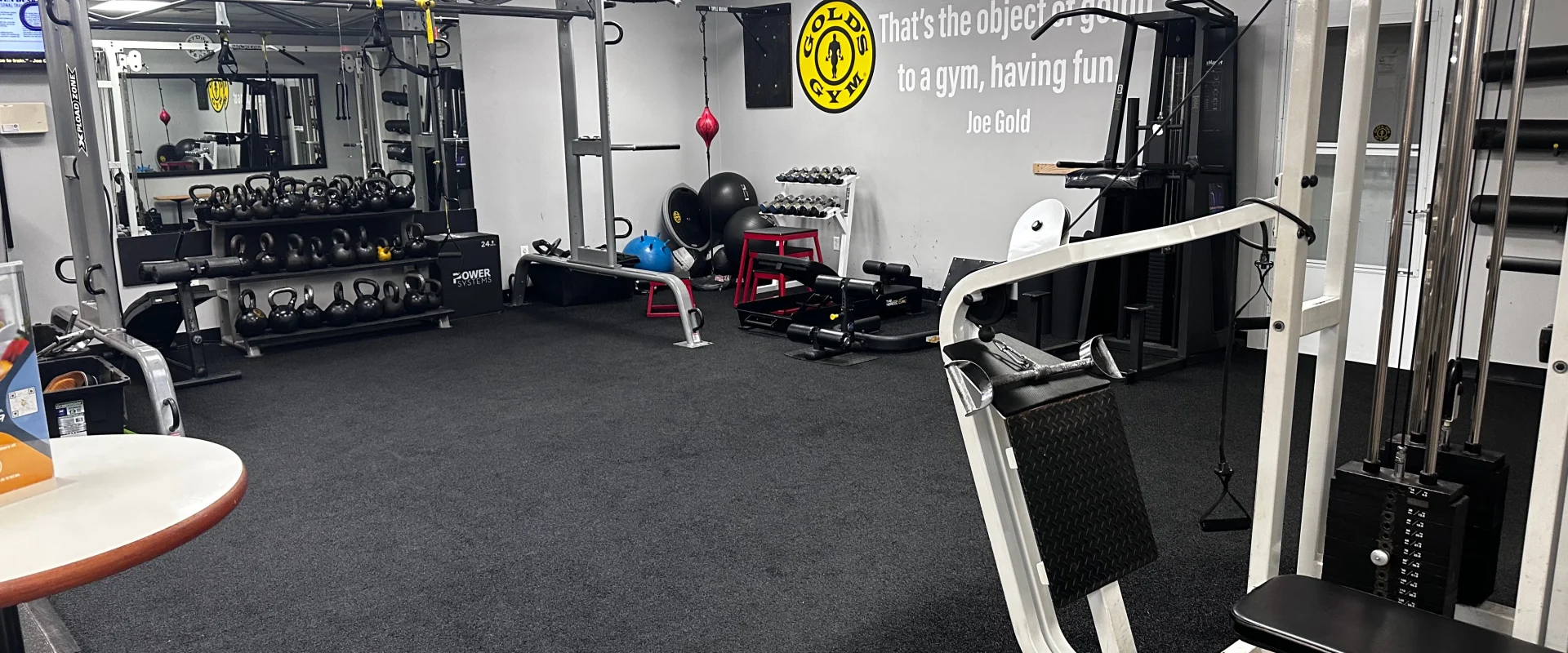 Functional training area at Gold’s Gym New Jersey, Bridgewater, featuring a black rubberized floor with a variety of functional training equipment. A multi-station functional rig with suspension training straps and pull-up bars is positioned against the mirrored wall. A rack of kettlebells is neatly arranged below, alongside medicine balls and stability balls. A cable machine is on the right, offering versatile exercise options. The wall features a motivational quote from Joe Gold, reinforcing the gym’s commitment to fitness. Various workout benches and small exercise tools like resistance bands and foam rollers are stored in the background.