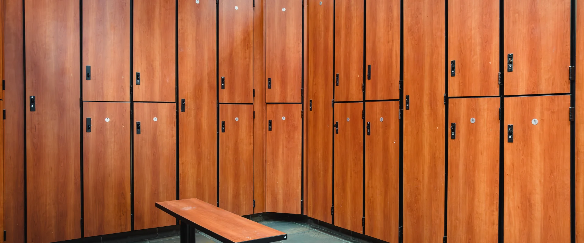 Locker room at Gold's Gym Waco featuring wood-paneled lockers with black accents, a matching wood bench, and a tiled floor with a natural stone look.
