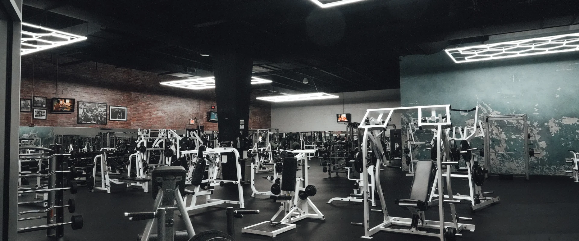 A spacious strength training area at Gold’s Gym Randleman with white resistance machines arranged across the floor, under geometric LED ceiling lighting, and surrounded by brick and distressed teal accent walls.