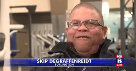 The image shows an older man with glasses and a serious expression, standing in what appears to be a news studio or interview setting, with a news banner visible in the background.